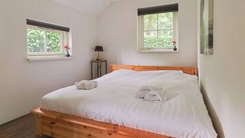 Ein Schlafzimmer mit einem hölzernen Bett, zwei Fenstern mit Blick auf Grünflächen, einem Nachttisch mit Lampe und einem an der Wand hängenden Gemälde.