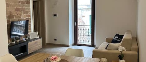 Un salón acogedor con techo de ladrillo, una mesa de comedor de madera, sillas blancas, un sofá color beige y un televisor sobre un mueble negro.