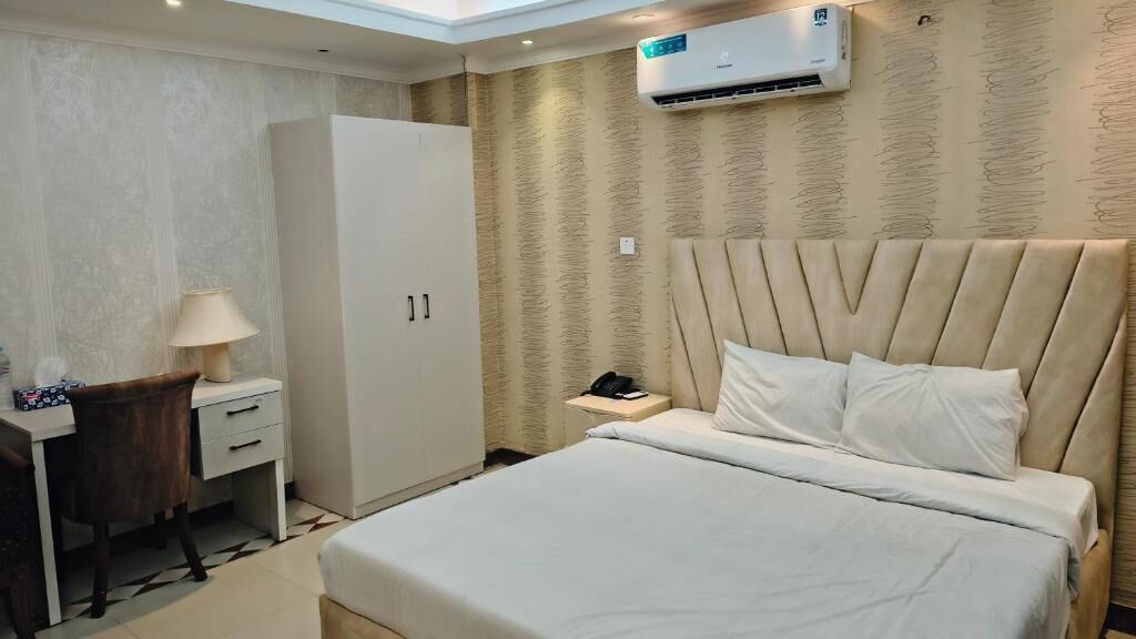A hotel room with a bed, bedside table, lamp, wardrobe, and an air conditioning unit.