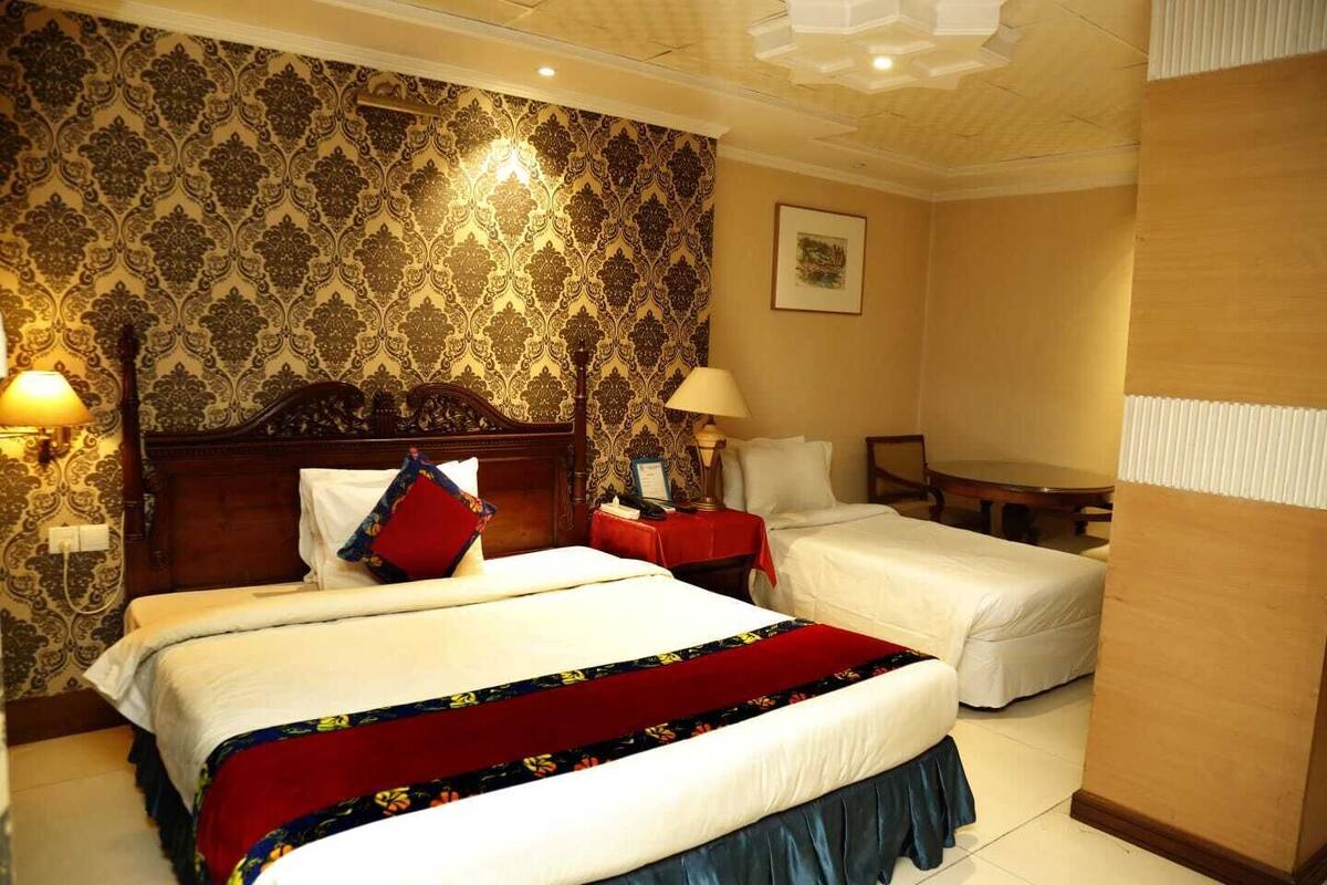 Ein Hotelzimmer mit zwei Betten, einem Holzkopfstück, einer roten und blauen Bettdecke, einem Nachttisch mit Lampe, einem gerahmten Bild an der Wand und einem Holztisch.