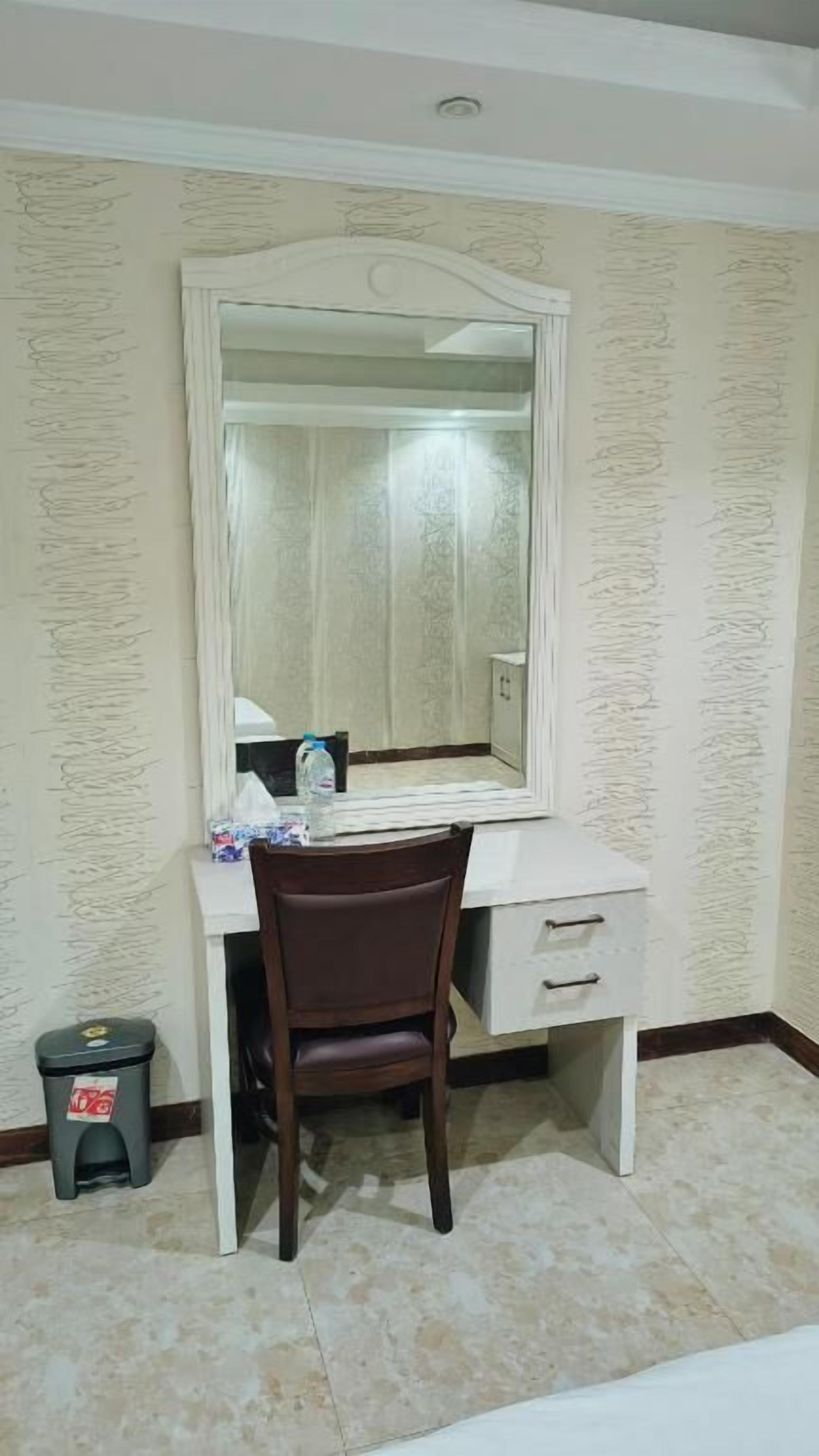 Una habitación con un escritorio blanco, un espejo grande, una silla marrón, un cubo de basura pequeño y un papel tapiz estampado.