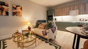 Un soggiorno moderno con divano, tavolino e lampada, una zona pranzo con tavolo e sedie, e una cucina con pensili in legno ed elettrodomestici.