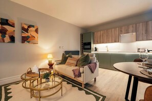 Een moderne woonkamer met een bank, salontafel en lamp, een eetgedeelte met tafel en stoelen, en een keuken met houten kastjes en apparatuur.