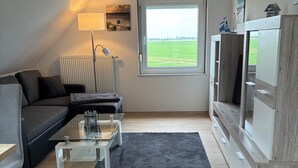 A modern living room with a sofa, coffee table, and TV stand, featuring a large window and a radiator. - Huus Diekblick Ditzum (Bunde)