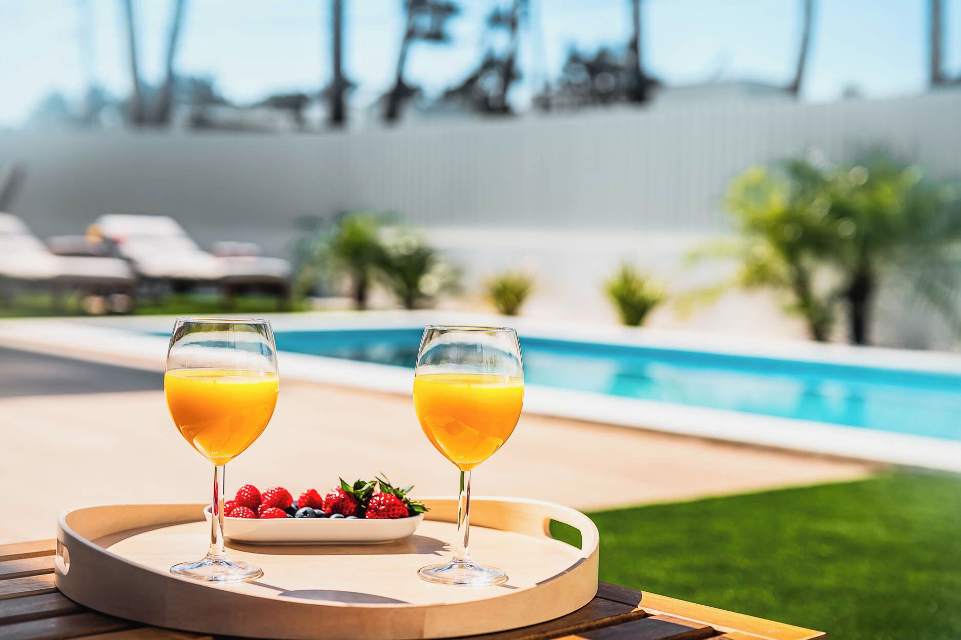 Two glasses of orange juice on a tray with strawberries and blueberries, by a pool with lounge chairs and palm trees in the background.