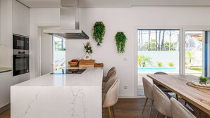 A modern kitchen with built-in appliances, a marble island, and a dining area with a view of a swimming pool. - OCEANBREEZE AROEIRA - Optional heated pool - Just 5 minutes from the beach (Aroeira)