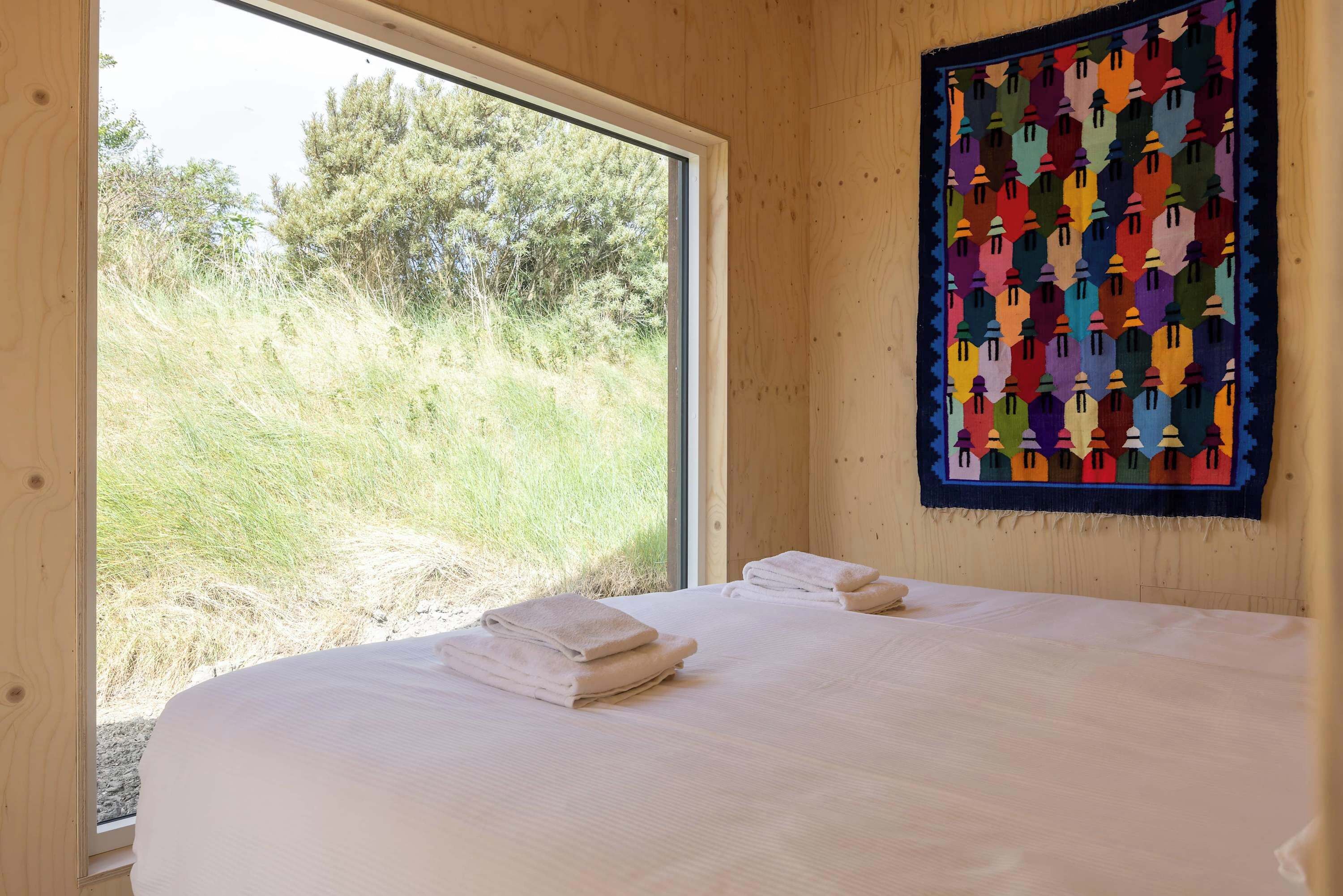 Una habitación con un amplio ventanal, una cama con toallas blancas ordenadas y un tapiz abstracto y colorido.