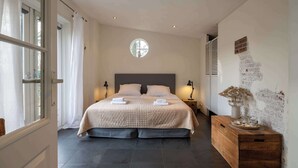 Ein Schlafzimmer mit einem großen Bett, einem Nachttisch mit einer Vase, einem Fenster mit Vorhängen und einer Wand mit Ziegelmuster.