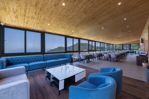 Amplio espacio interior con un techo de madera, grandes ventanales que ofrecen vistas a las montañas, un sofá azul y una mesa de centro blanca.