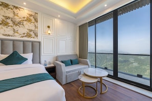 A hotel room with a large bed, a sofa, a small table, and a view of the landscape through large windows.