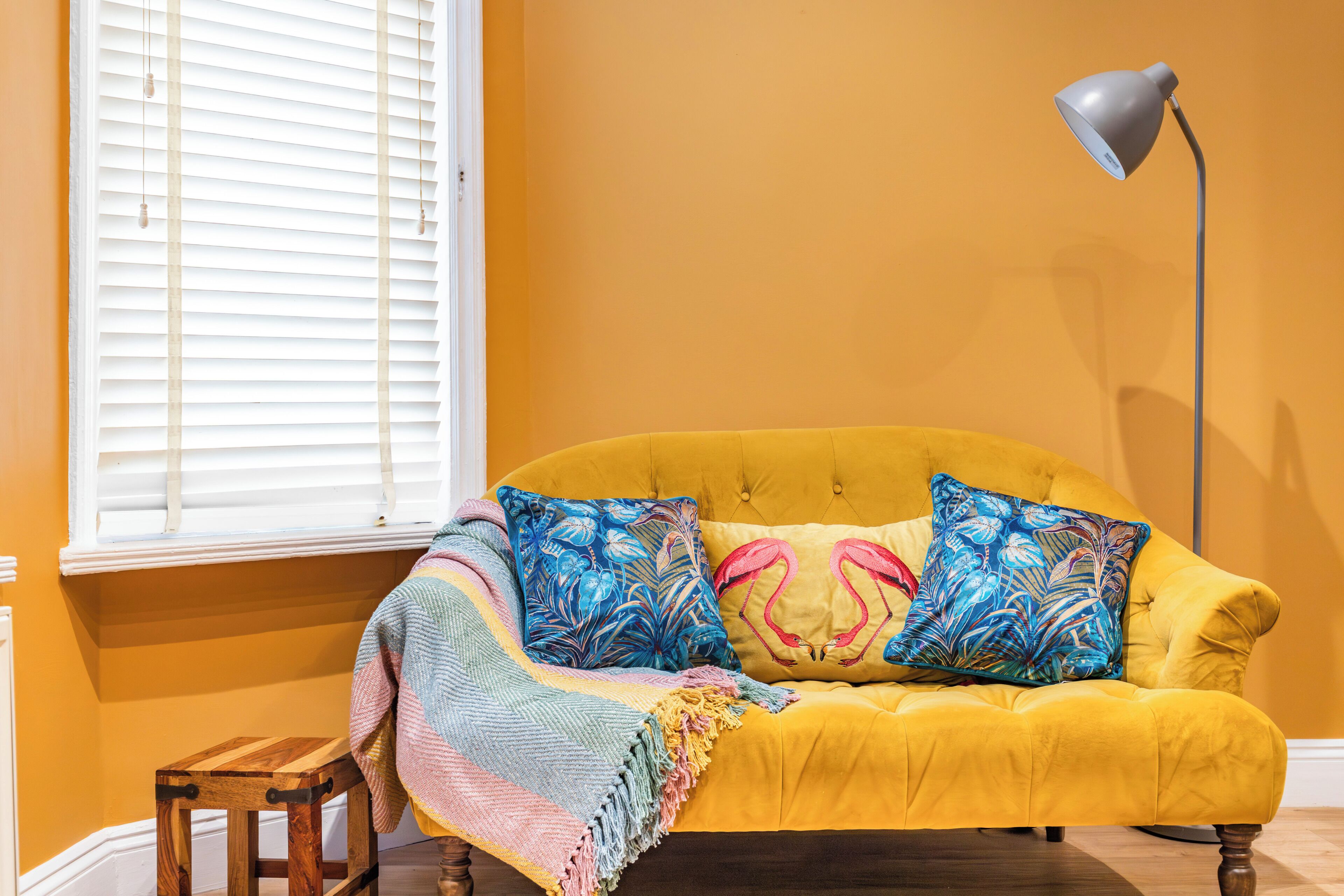Ein gelbes Sofa mit gemusterten Kissen, ein Holzschemel und eine Stehlampe in einem Raum mit orangefarbenen Wänden.