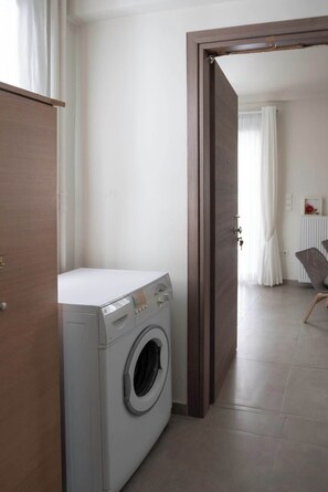 Een wasserette met een wasmachine, een houten deur en uitzicht op een woonruimte met een fauteuil en gordijnen.