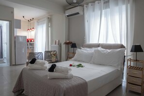 A neatly made bed with pillows and towels, a bedside table with a lamp, a chair, and a view of a kitchen area. - Central & Spacious Athenian Penthouse Getaway (Athina)