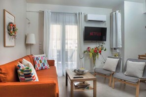 Ein Wohnzimmer mit einer Couch, Sesseln, einem Couchtisch, einem Fernseher und einem Fenster mit Vorhängen.