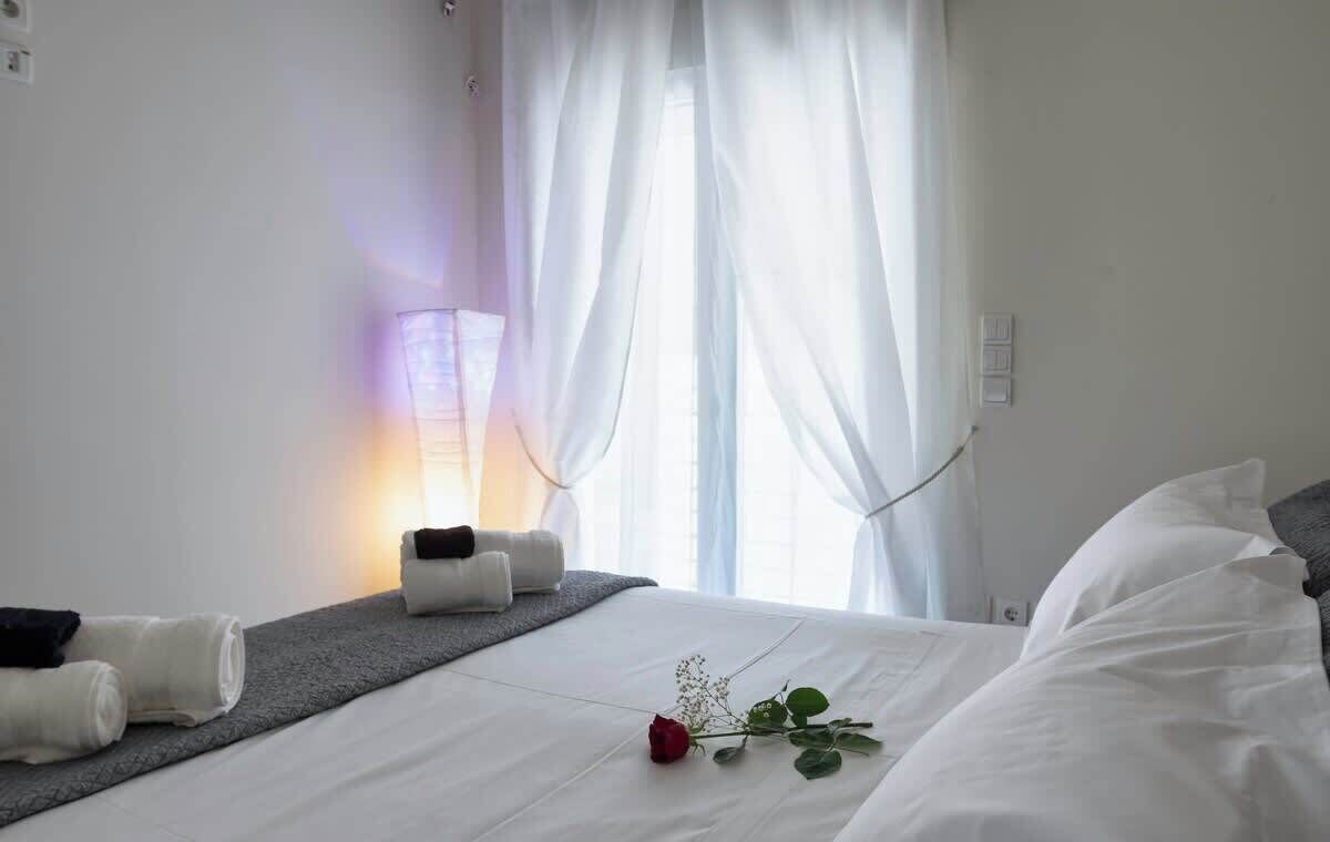 Et hotelværelse med en seng, et natbord med håndklæder, en lampe og et vindue med let gardin.