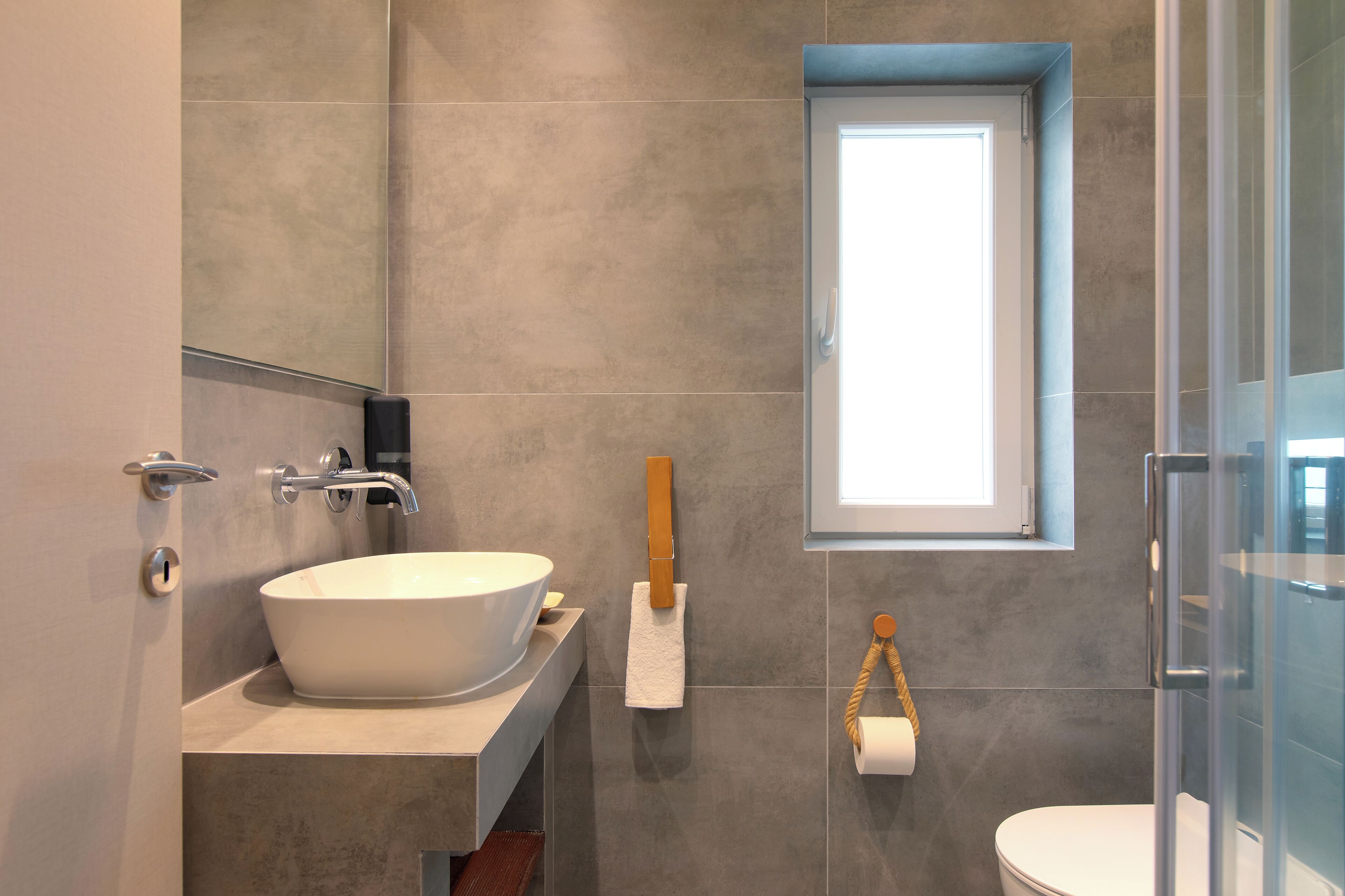 Un baño moderno con un lavamanos grande tipo recipiente, un perchero, un inodoro y una ventana pequeña.