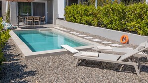 A swimming pool with lounge chairs and an umbrella, surrounded by gravel and greenery. - KAELA Pool Villas (Agios Nikolaos)