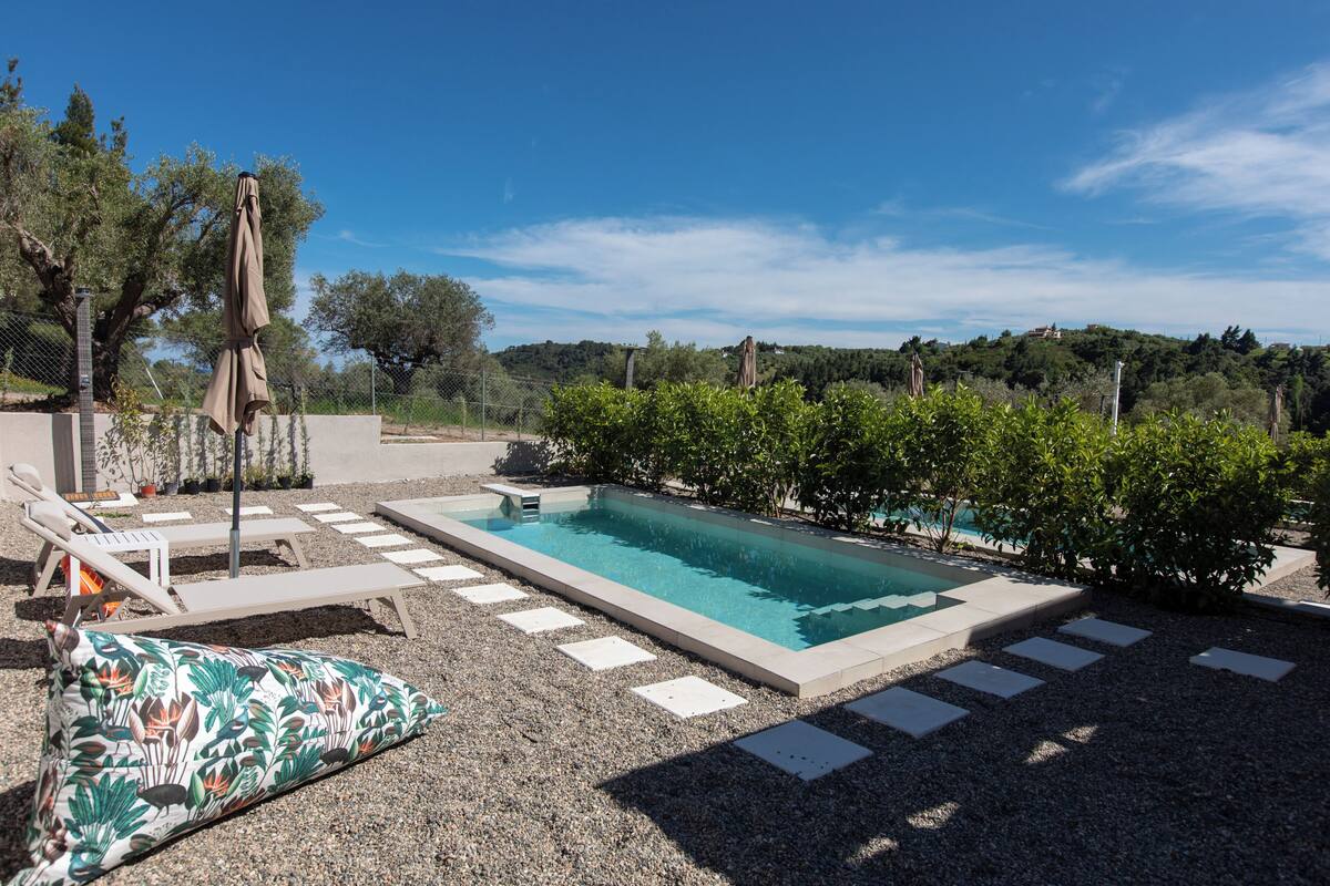 Una piscina rectangular con superficie de agua azul cristalina, rodeada por una zona de grava con piedras que conducen a tumbonas y una sombrilla.