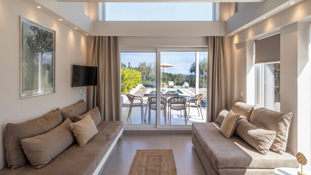 A spacious living area with two sofas, a flat-screen TV, and a large sliding glass door leading to an outdoor patio with a pool. - KAELA Pool Villas (Agios Nikolaos)