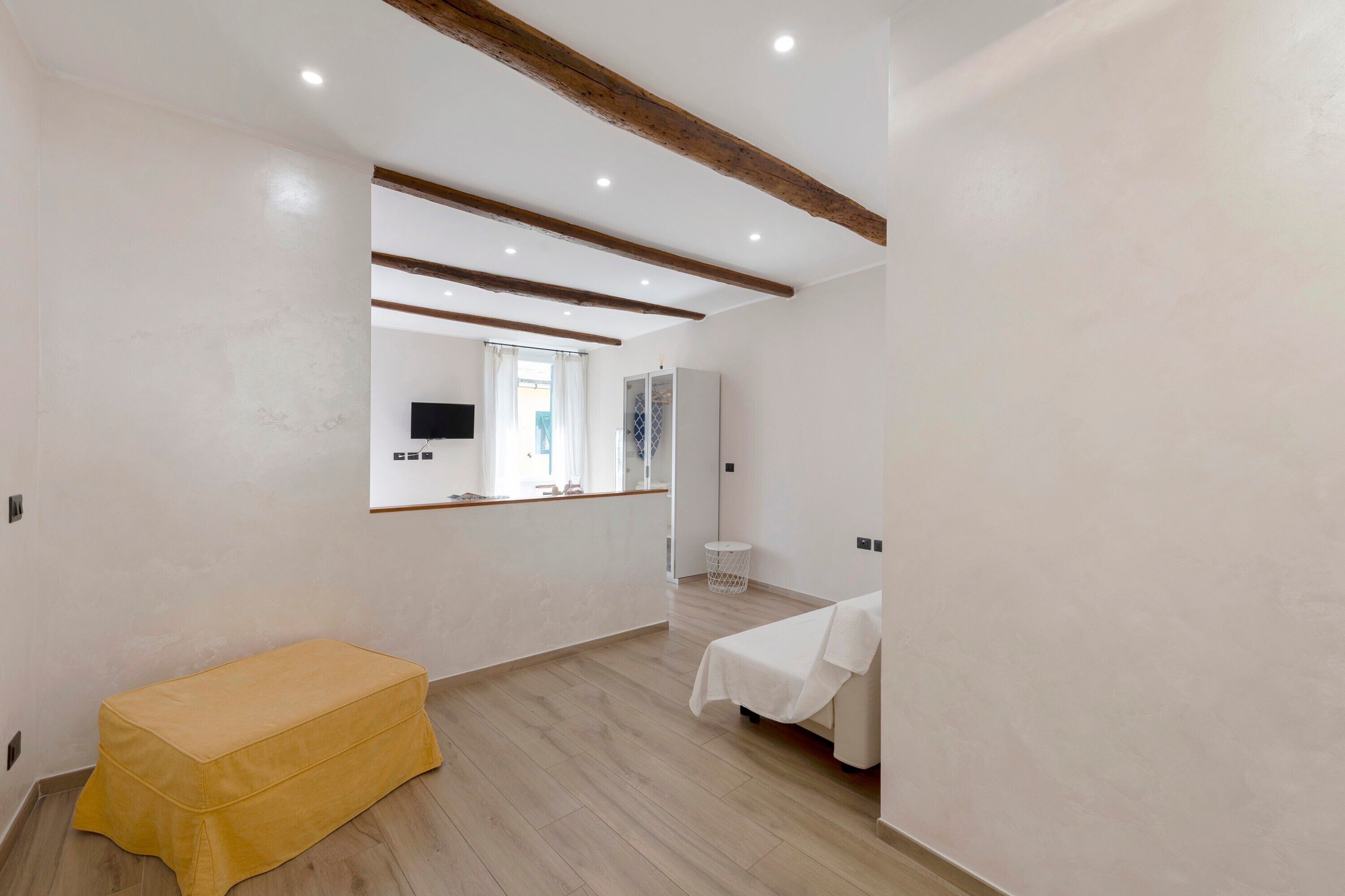 A room with a wooden bench, a bed with a white cover, a mirror, and a television on the wall.