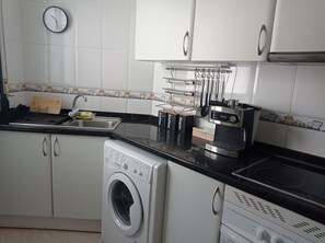 A kitchen with a washing machine, coffee machine, and a clock on the wall. - Edificio Azul V Dúplex 2º (Playa de Gandía)