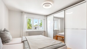 Ein Schlafzimmer mit Bett, Fenster, Heizkörper, Spiegel und einer Holzbank.