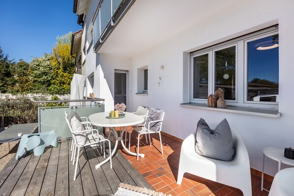 Eine Terrasse mit einem runden Tisch und Stühlen, einer weißen Wand und einem Fenster mit Blick auf einen Garten.