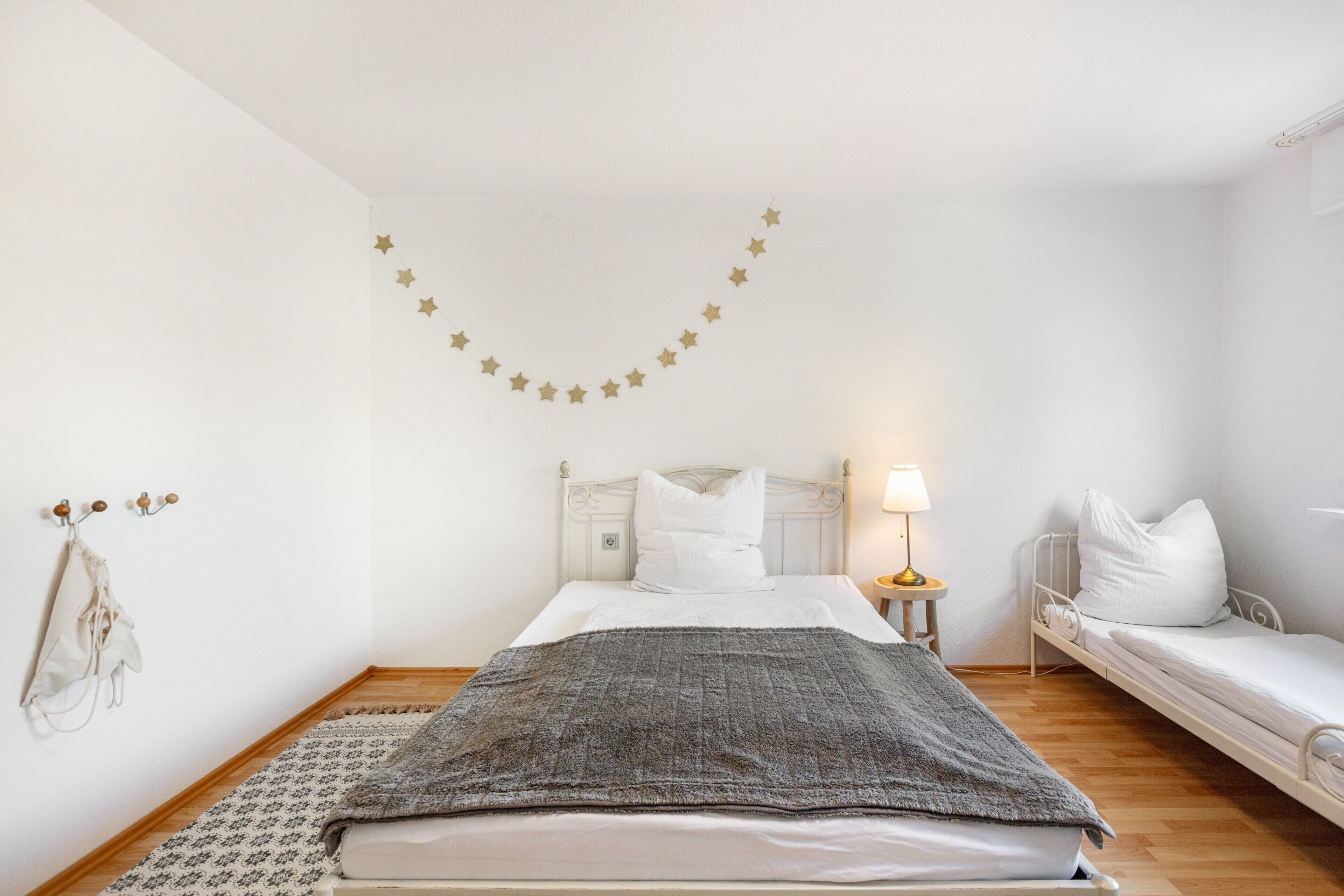 A bedroom with a bed, a bedside table with a lamp, a smaller bed, a wall-mounted star decoration, and a coat hanging on the wall.