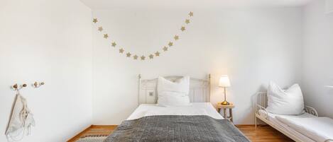A bedroom with a bed, a bedside table with a lamp, a smaller bed, a wall-mounted star decoration, and a coat hanging on the wall.