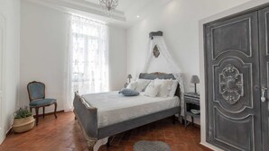 Een slaapkamer met een bed, een stoel, een nachtkastje, een lamp, een raam met gordijnen, een kroonluchter en een deur.
