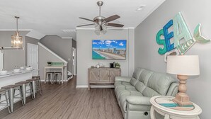 A modern living room with a sofa, a dining area with stools, a ceiling fan, and a wall-mounted TV. - 7 Bedrooms-OceanLakes-Sleeps18-GolfCart Included (Myrtle Beach)