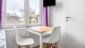 Una habitación con una mesa y sillas blancas, un televisor montado en la pared y un ventanal con cortinas moradas.