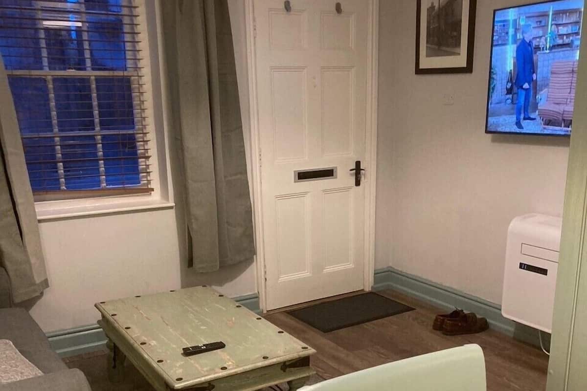 A room with a white door, a small table, a chair, a sofa, a television, and a window with blinds.