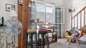 Une pièce avec un grand ventilateur, deux tabourets, une chaise, une fenêtre avec des rideaux et un escalier.