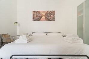Una cama bien hecha con ropa y toallas blancas, una silla, una lámpara de pie y un cuadro con una escena de un canal en la pared.