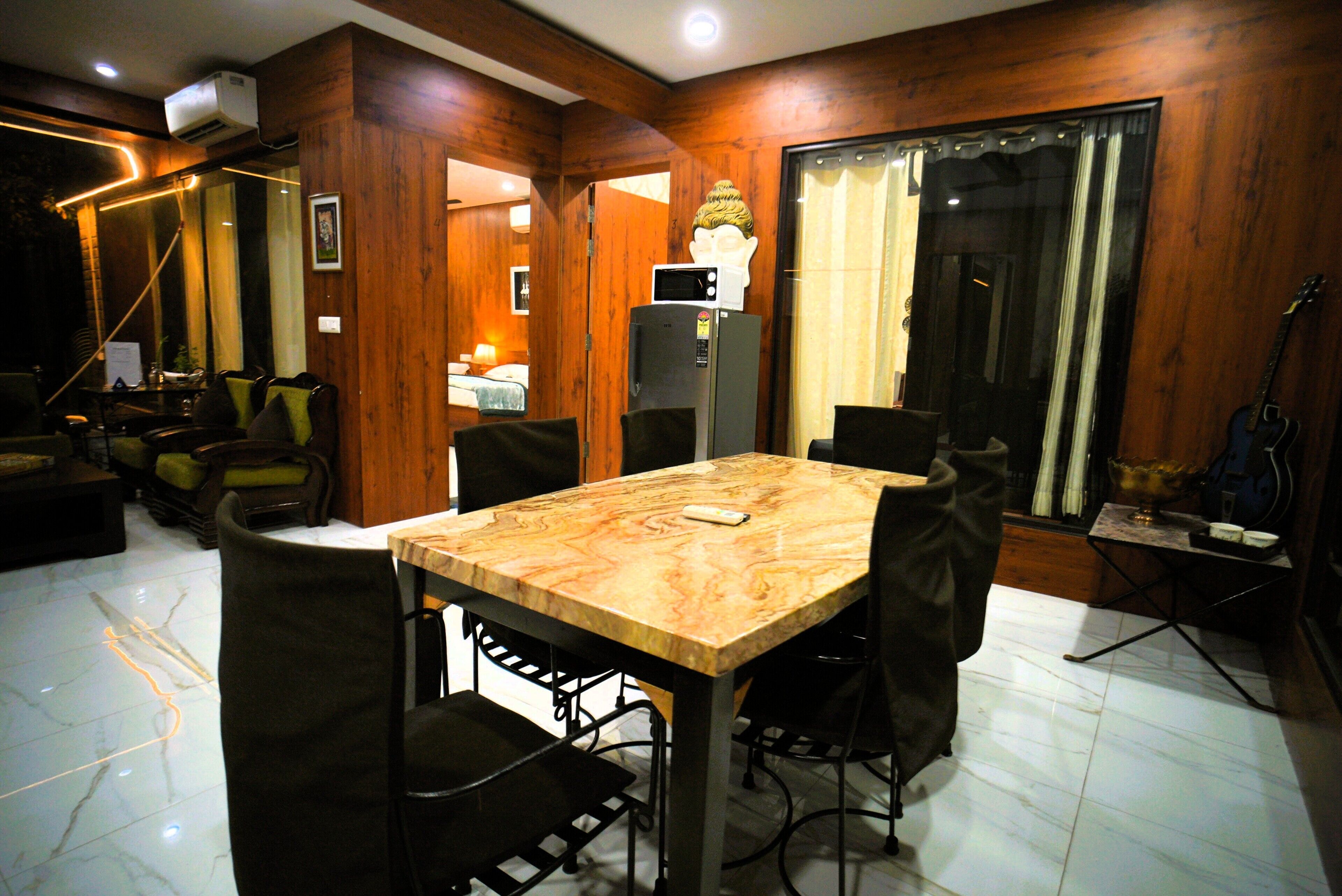Una habitación con pared de paneles de madera, una mesa de comedor con sillas, un refrigerador, una guitarra y un ventanal con cortinas.