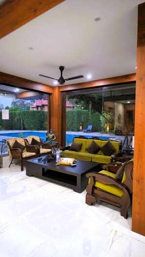 A modern living room with a pool view, wooden ceiling beams, and a ceiling fan. - The Mannat Farms (Taoru)