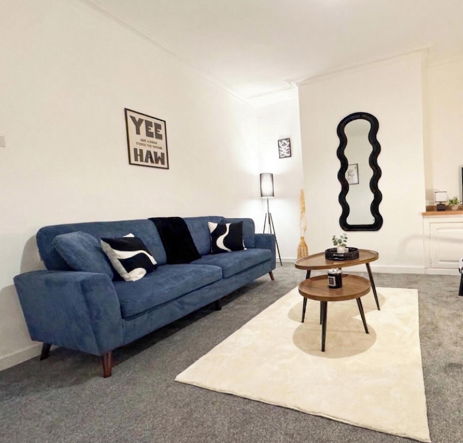 A living room with a blue sofa, a round wooden coffee table, a white rug, and a wall-mounted mirror.