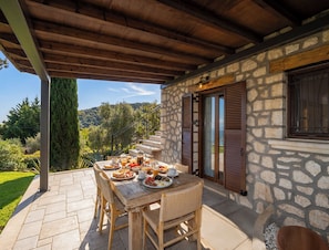 A covered patio with a stone wall, wooden furniture, and a table set with breakfast. - Anemones Villas (Lefkada)