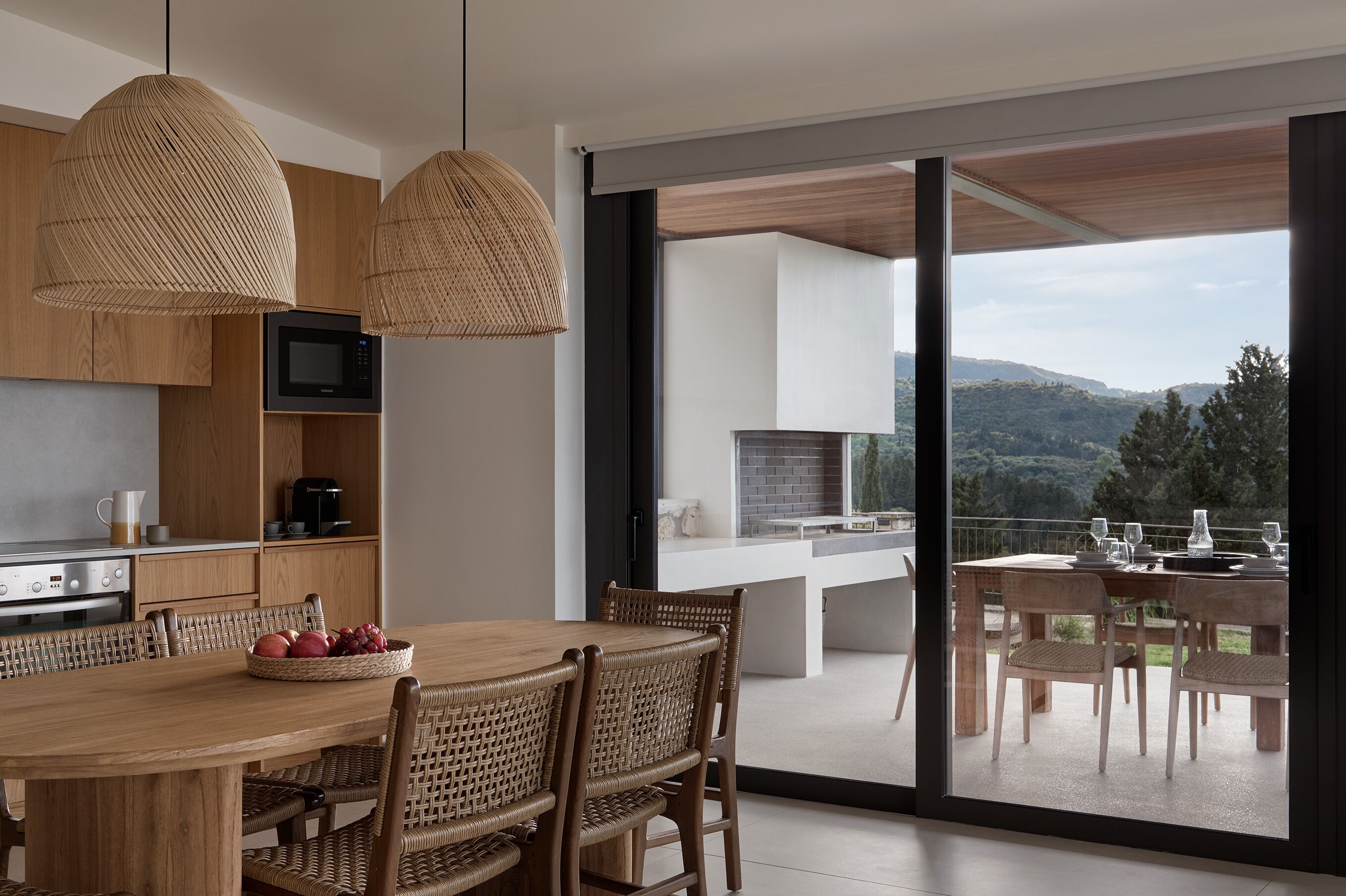 A modern kitchen with wooden cabinets, a dining area with wicker chairs, and a view of a balcony with a table set for dining.