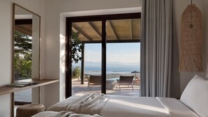 A bedroom with a large bed, a view of the outdoors, a hanging light fixture, and a shelf with a book. - Anemones Villas (Lefkada)
