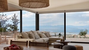 Ein modernes Wohnzimmer mit großem Fenster, einer Couch, einem Esstisch mit Obstschale und Korbmöbeln.