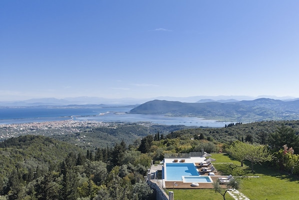 A pool with lounge chairs, a view of a coastal town, and a mountainous landscape. - Anemones Villas (Lefkada)