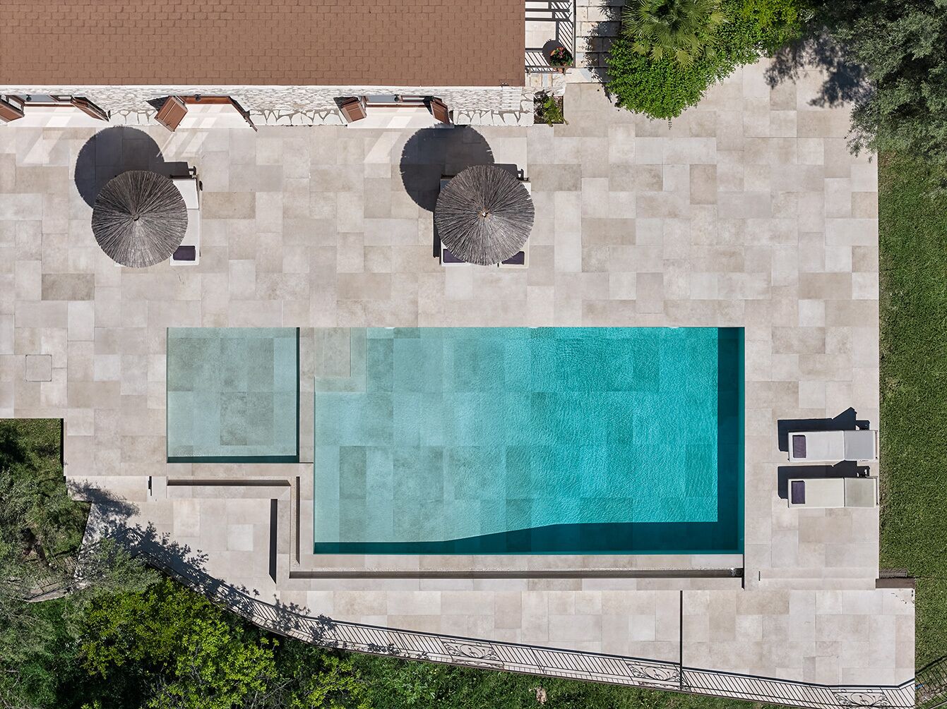 An aerial view of a swimming pool with adjacent lounge areas, surrounded by a paved deck and greenery.