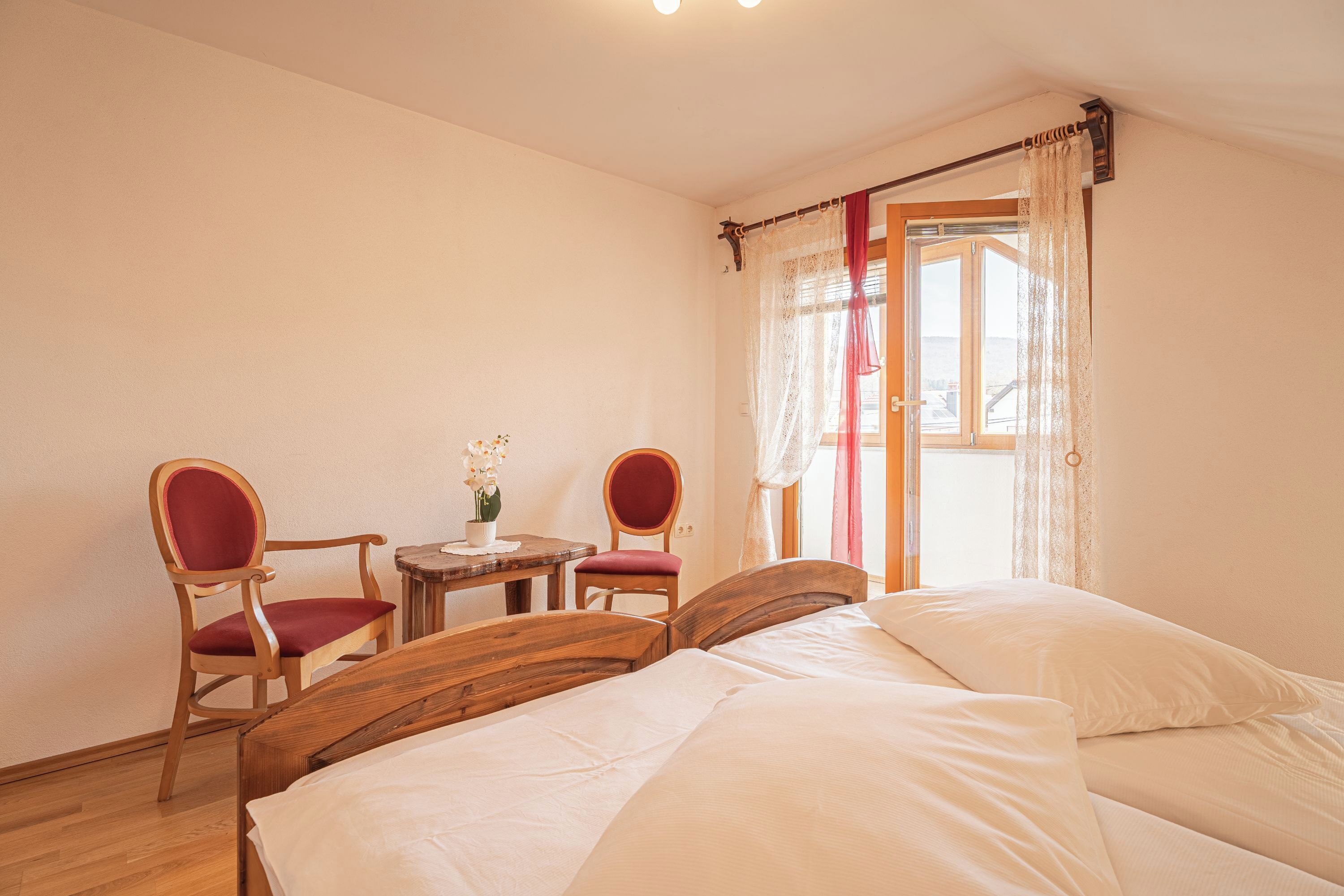 Habitació amb un llit, dues cadires, una taula petita amb un vas i una finestra amb cortines.