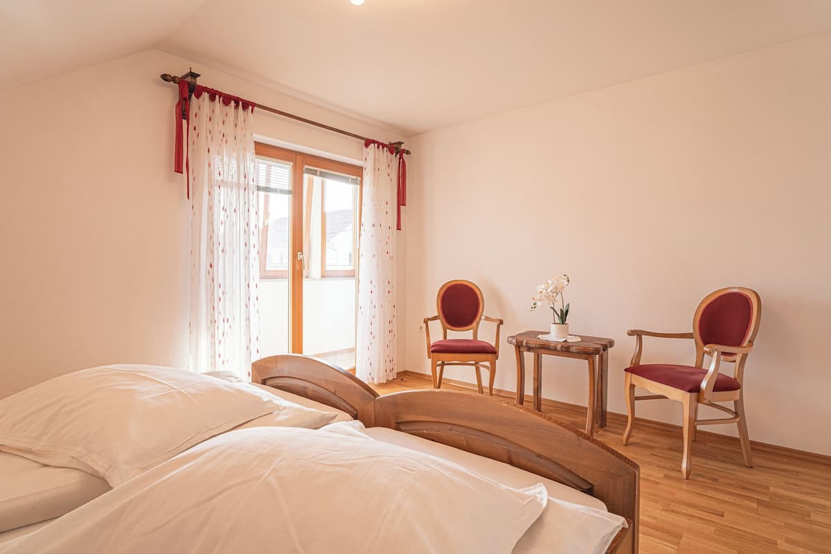 Un dormitorio con una cama, dos sillas, una mesita con un florero y una ventana con cortinas.