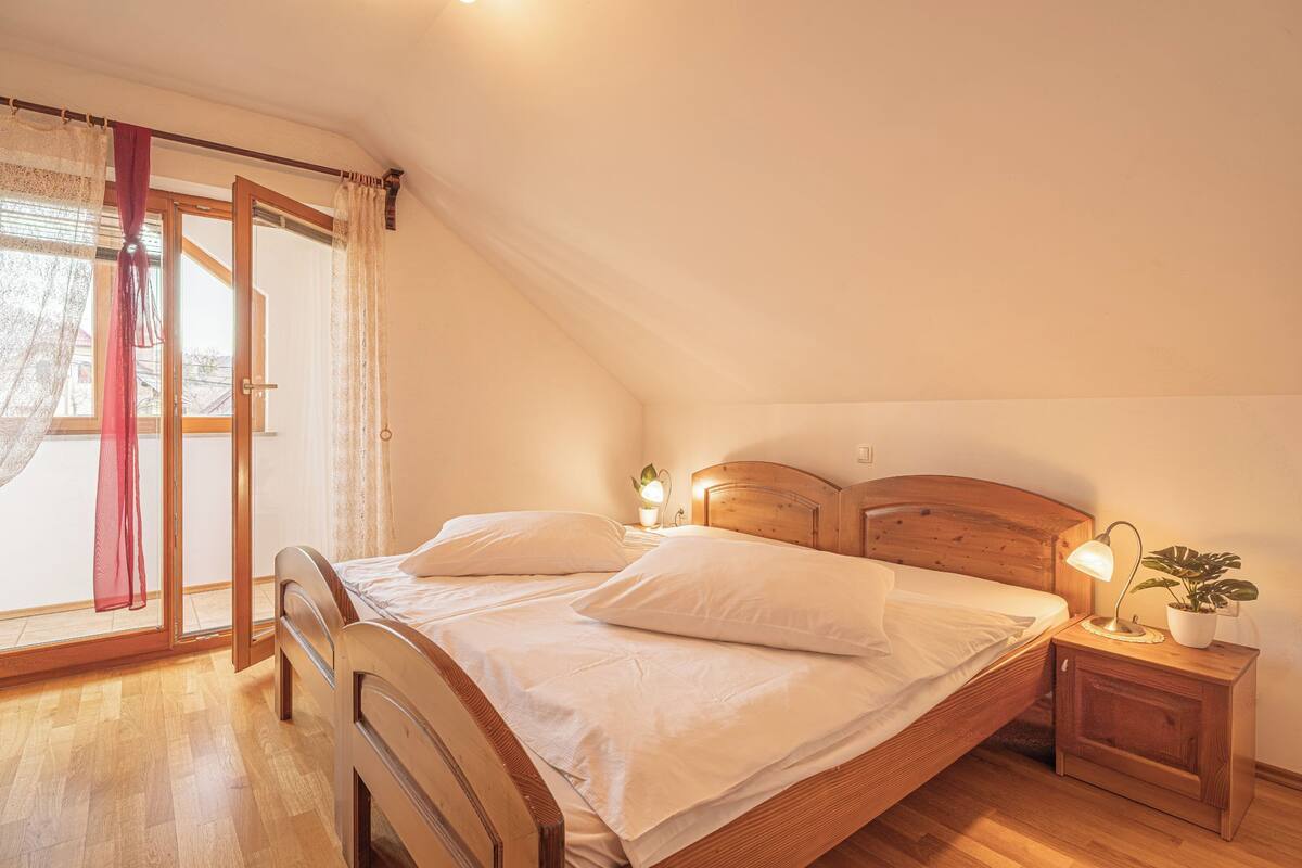 Un dormitorio con una cama de madera, una mesita de noche con lámpara, una planta en maceta y un ventanal corredizo con cortinas.