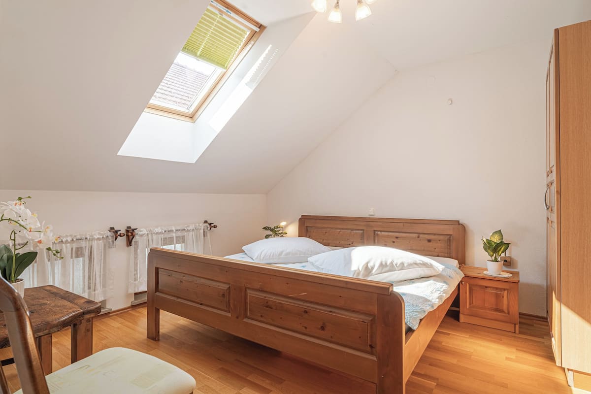Un dormitorio con cama de madera, una mesita de noche pequeña, una silla, una ventana con vistas y una lámpara en el techo.