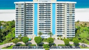 Un hôtel à plusieurs étages, avec une bande bleue verticale au centre, situé près d’une plage, avec un parking visible et de la végétation.