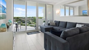 Un salon moderne avec un canapé d’angle gris, une grande fenêtre donnant sur un paysage verdoyant et une porte-fenêtre coulissante qui mène à un balcon extérieur.